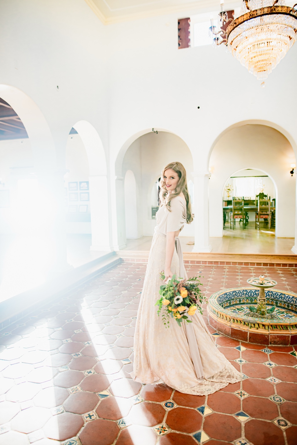 Woman in a cream dress holding flowers in a sunlit tiled courtyard